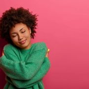 Cheerful young romantic woman expresses self love and care, tilts head and smiles gently, wears green oversized jumper, embraces own body, closes eyes, stands in studio against pink background