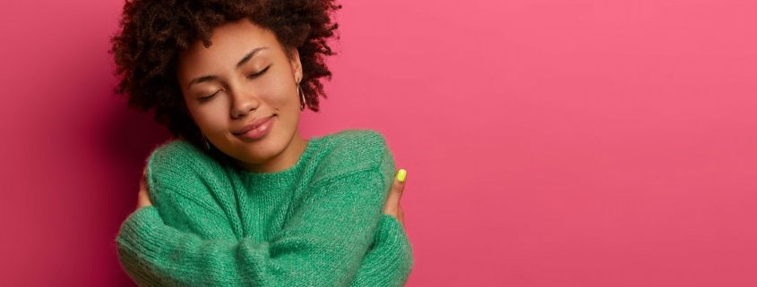 Cheerful young romantic woman expresses self love and care, tilts head and smiles gently, wears green oversized jumper, embraces own body, closes eyes, stands in studio against pink background