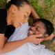 Two black people lying on the grass together, smiling and looking lovingly at each other. One has their hand on the cheek of the other, gently holding their face.
