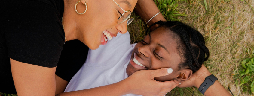 Two black people lying on the grass together, smiling and looking lovingly at each other. One has their hand on the cheek of the other, gently holding their face.
